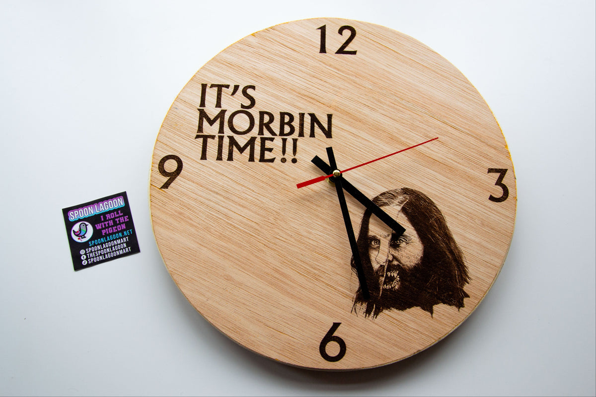 A round wooden wall clock with a natural light wood finish, featuring the engraved text 'IT'S MORBIN TIME!!' and a portrait engraving near the 6 o'clock position. The clock has black Arabic numerals and a red second hand, and is shown on a plain white background next to a small sticker.
